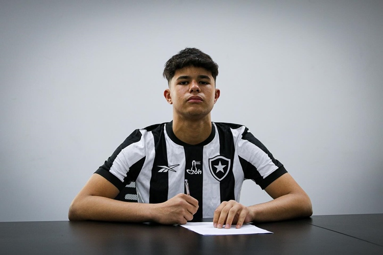 Bruno Samudio está sentado em uma mesa, vestindo uma camisa listrada em preto e branco do Botafogo. Ele segura uma caneta e parece estar prestes a assinar um documento. O ambiente é simples, com uma parede branca ao fundo, sugerindo um escritório ou sala de reuniões. A expressão do jovem é séria e concentrada, indicando a importância do momento. A cena captura um instante de compromisso e expectativa.