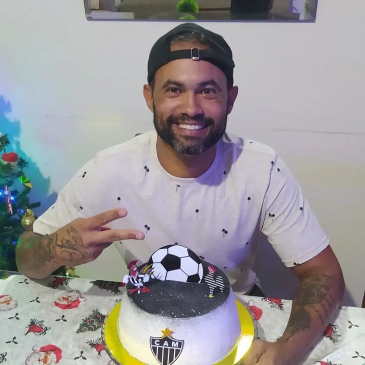 Na foto, o goleiro Bruno sorridente está sentado à mesa com um bolo decorado com tema de futebol e o escudo do Atlético Mineiro. Ele usa uma camiseta clara com estampas pequenas e um boné virado para trás. O homem faz um sinal de paz com os dedos. Ao fundo, há uma árvore de Natal decorada, sugerindo uma celebração festiva. A cena transmite alegria e entusiasmo, destacando a paixão pelo esporte.