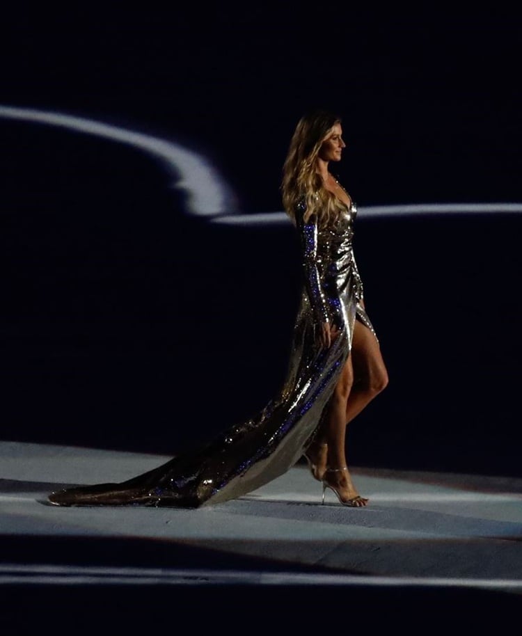 A imagem apresenta uma mulher usando um vestido de festa deslumbrante, prateado e brilhante. O vestido é longo, com uma cauda elegante que se arrasta pelo chão. Possui um corte assimétrico, destacando uma fenda alta que revela a perna. As mangas longas e o tecido cintilante refletem a luz, criando um efeito glamouroso.