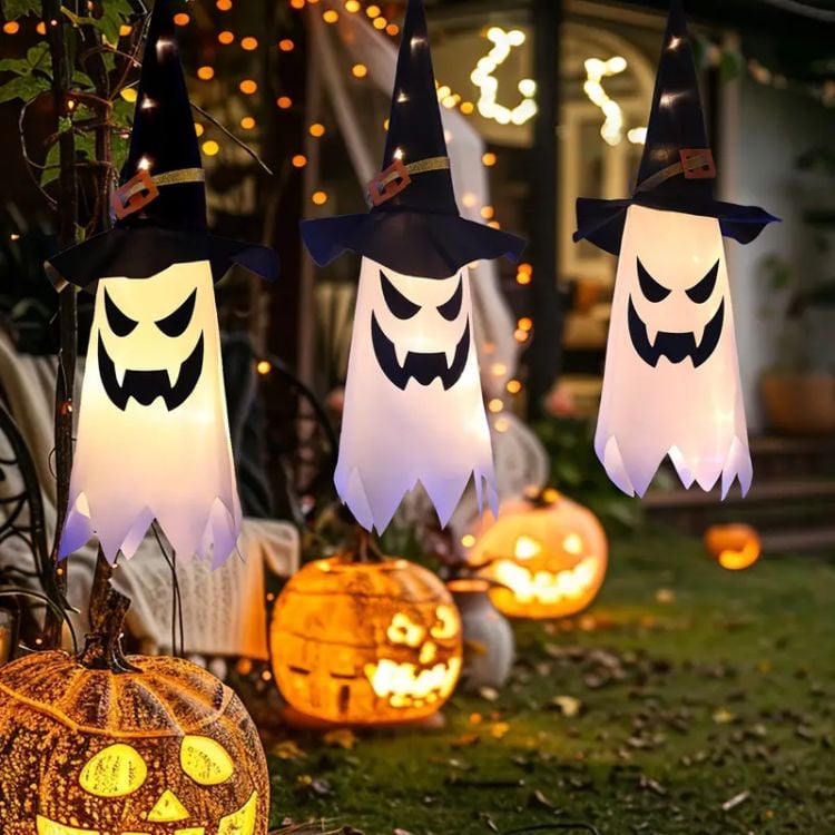 A imagem mostra uma decoração de Halloween ao ar livre com três fantasmas suspensos. Eles têm rostos assustadores com bocas abertas e olhos angulares, além de usarem chapéus de bruxa pretos com detalhes dourados. Ao fundo, há várias abóboras iluminadas esculpidas com expressões assustadoras, criando um ambiente típico de Halloween.