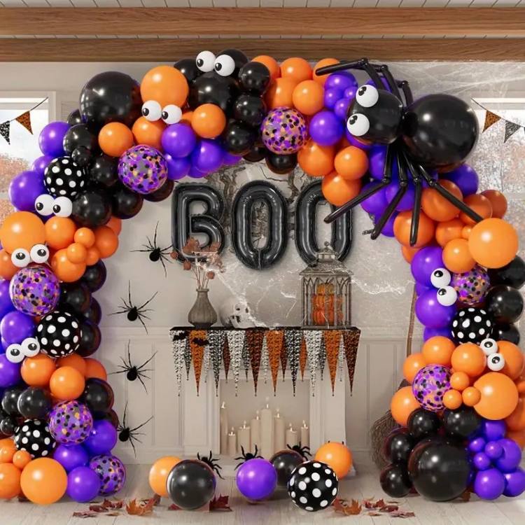 A imagem mostra uma decoração de Halloween vibrante e divertida, composta por um arco de balões nas cores preto, laranja, roxo e alguns com padrões de bolinhas. O arco é adornado com olhos grandes, representando aranhas e outros elementos temáticos. No topo do arco, há uma grande aranha feita de balões pretos. Atrás do arco, na parede, estão balões metálicos prateados formando a palavra "BOO". A mesa decorada logo abaixo tem uma toalha de franjas em núcleos temáticos de Halloween e está cercada por velas e pequenas aranhas decorativas.