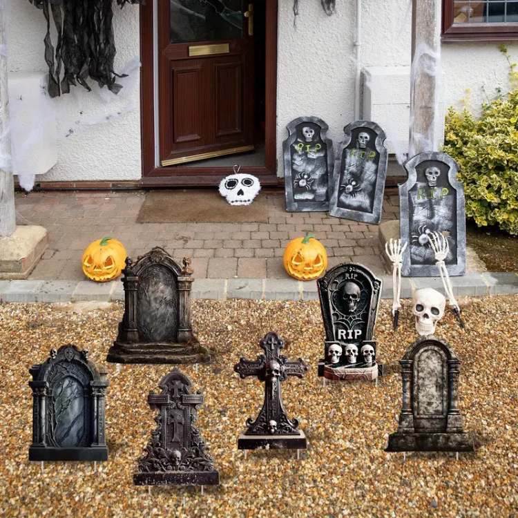 A imagem mostra uma decoração de Halloween na entrada de uma casa, com um tema de cemitério assustador. Várias lápides estão dispostas no chão de pedras, com designs antigos e góticos, algumas com a inscrição "RIP" e caveiras esculpidas. Ao redor, há abóboras esculpidas (jack-o'-lanterns) iluminadas, adicionando um toque tradicional de Halloween. Perto da porta, há esqueletos e braços saindo do solo, criando uma ilusão de mortos-vivos emergindo da terra. O ambiente é complementado com teias de aranha falsas suspensas nas paredes, e a porta de entrada está decorada com mais elementos sombrios.