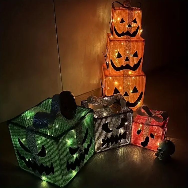 A imagem mostra uma decoração temática de Halloween composta por caixas iluminadas com rostos de abóboras esculpidos no estilo "jack-o'-lantern." As caixas estão organizadas em dois grupos: à direita, três caixas empilhadas em tamanhos decrescentes com luzes internas laranja, e à esquerda, três caixas menores e separadas, iluminadas em tons de verde, branco e vermelho. Cada caixa tem expressões faciais assustadoras e são envolvidas por laços decorativos. Ao lado das caixas, há uma pequena caveira, que adiciona um toque extra ao clima sombrio e festivo da cena.