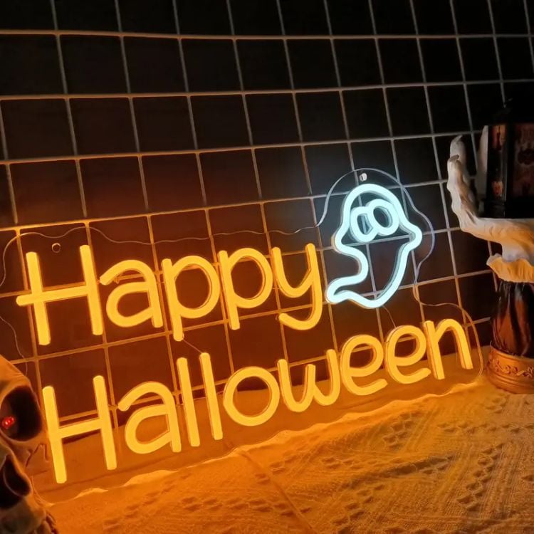 A imagem mostra uma placa de neon com a mensagem "Happy Halloween" em letras laranja brilhantes. Ao lado da palavra "Halloween," há o desenho de um pequeno fantasma estilizado, iluminado em luz branca. A placa está posicionada contra um fundo de azulejos pretos e repousa sobre uma superfície com um tecido claro. À esquerda, é possível ver parte de uma caveira, e à direita, parte de uma decoração, provavelmente relacionada ao tema de Halloween, com uma mão esquelética segurando um objeto. A iluminação suave e o estilo neon dão um clima festivo e moderno à decoração.