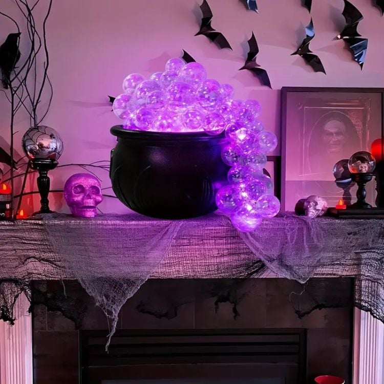 A imagem apresenta uma decoração de Halloween com um caldeirão preto no centro, transbordando com esferas translúcidas iluminadas por luzes roxas, criando o efeito de "poção mágica" ou vapor saindo do caldeirão. Ao redor, há vários elementos decorativos típicos do tema: uma caveira roxa brilhante à esquerda, uma bola de cristal em um suporte, velas vermelhas e um quadro com uma imagem assustadora de um rosto. Na parede ao fundo, várias figuras de morcegos negros estão penduradas, contribuindo para o clima sombrio. A lareira está coberta com um tecido envelhecido, realçando o estilo assustador da decoração.