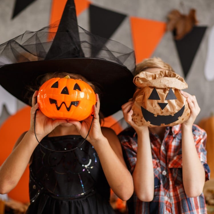 Duas crianças de pele clara em festa de Halloween. A da direita usa fantasia de bruxa e bolsa de abóbora no rosto e outra de camisa xadrez e saco de pão com rosto de abóbora