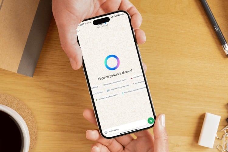 A imagem mostra uma mão segurando um smartphone com a tela aberta no WhatsApp. No centro, há um círculo colorido em tons de azul e rosa, representando a IA do Meta. Abaixo, lê-se "Faça perguntas à Meta AI". O fundo é uma mesa de madeira clara com um caderno, uma caneta e uma xícara de café.