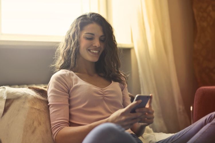 A imagem mostra uma mulher sorrindo enquanto usa seu smartphone, sentada confortavelmente em um sofá. Ela representa o uso de inteligência artificial na terapia, destacando a conveniência e acessibilidade do suporte emocional digital. A luz suave ao fundo cria um ambiente acolhedor e tranquilo, enfatizando bem-estar e inovação.