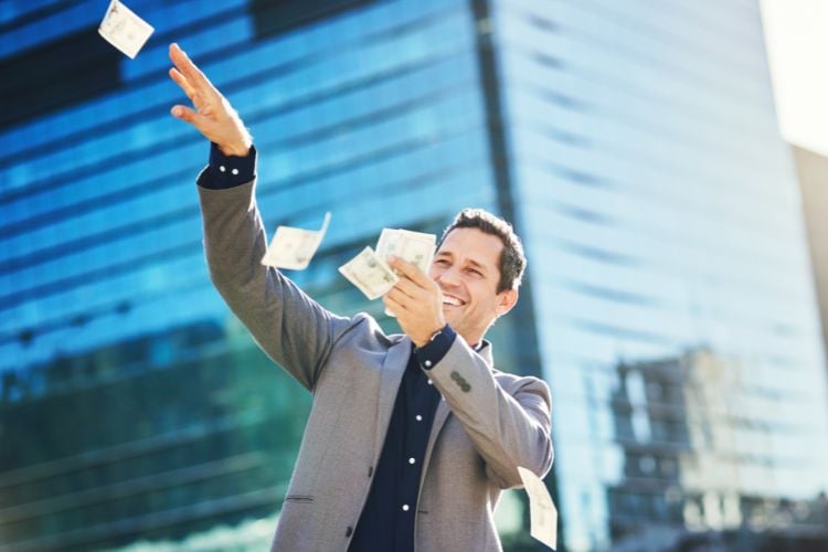 Um homem sorridente, vestindo um terno cinza, está jogando notas de dinheiro ao ar em frente a um prédio moderno. A cena transmite uma sensação de alegria e liberdade, como se ele tivesse acabado de ganhar na loteria ou feito um investimento bem-sucedido. A luz do sol reflete nos vidros do edifício, criando um ambiente vibrante.