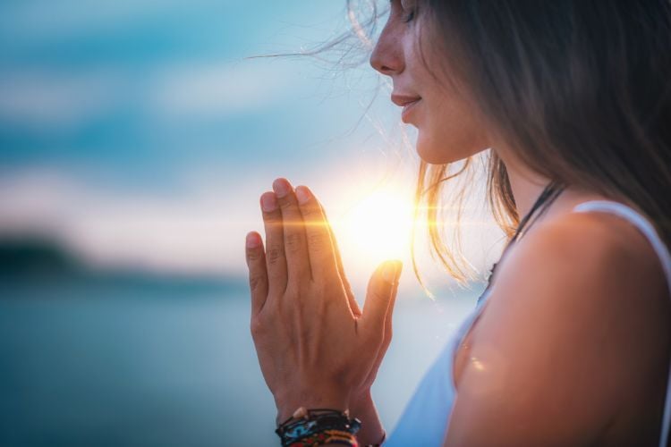 Mulher de pele clara usando regata branca, com mãos unidas em oração e nascer do sol ao fundo 