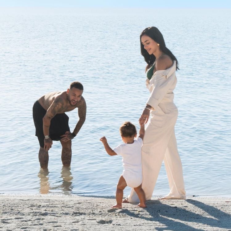 Foto de Neymar de bermuda preta abaixado no mar, olhando para Mavie de costas usando body branco e de mãos dadas com Bruna Biancardi, de look branco