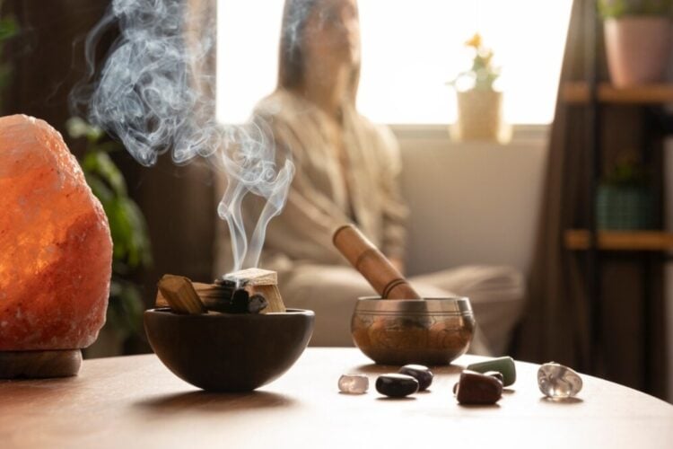 Foto de mesa com cristais e incenso e mulher de pele clara sentada meditando ao fundo desfocado