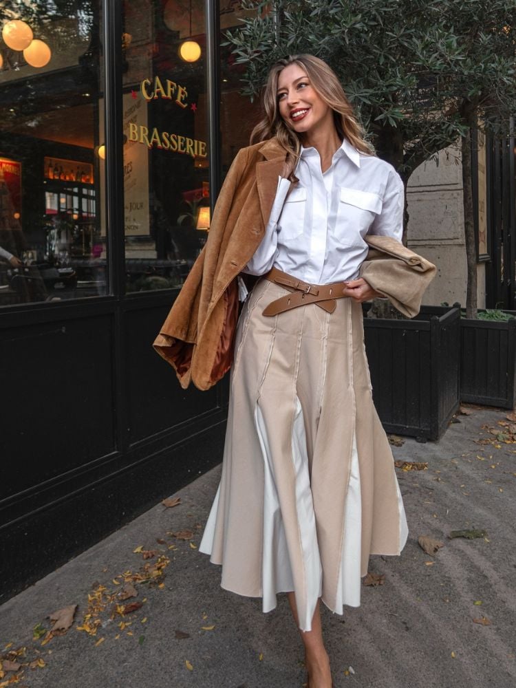 A imagem mostra uma mulher sorridente em frente a uma brasserie com estilo europeu. Ela está usando um conjunto elegante, composto por uma camisa branca de mangas longas com bolsos frontais, uma saia longa bege com detalhes plissados em branco, que cria uma aparência sofisticada e moderna. A saia é amarrada com um cinto marrom que realça a cintura. Ela também carrega um casaco de veludo marrom sobre um dos ombros, o que combina bem com o tom neutro do restante da roupa. O ambiente ao fundo, com folhas caídas no chão, sugere uma cena de outono em uma cidade urbana, com luzes aconchegantes refletindo de dentro do café.