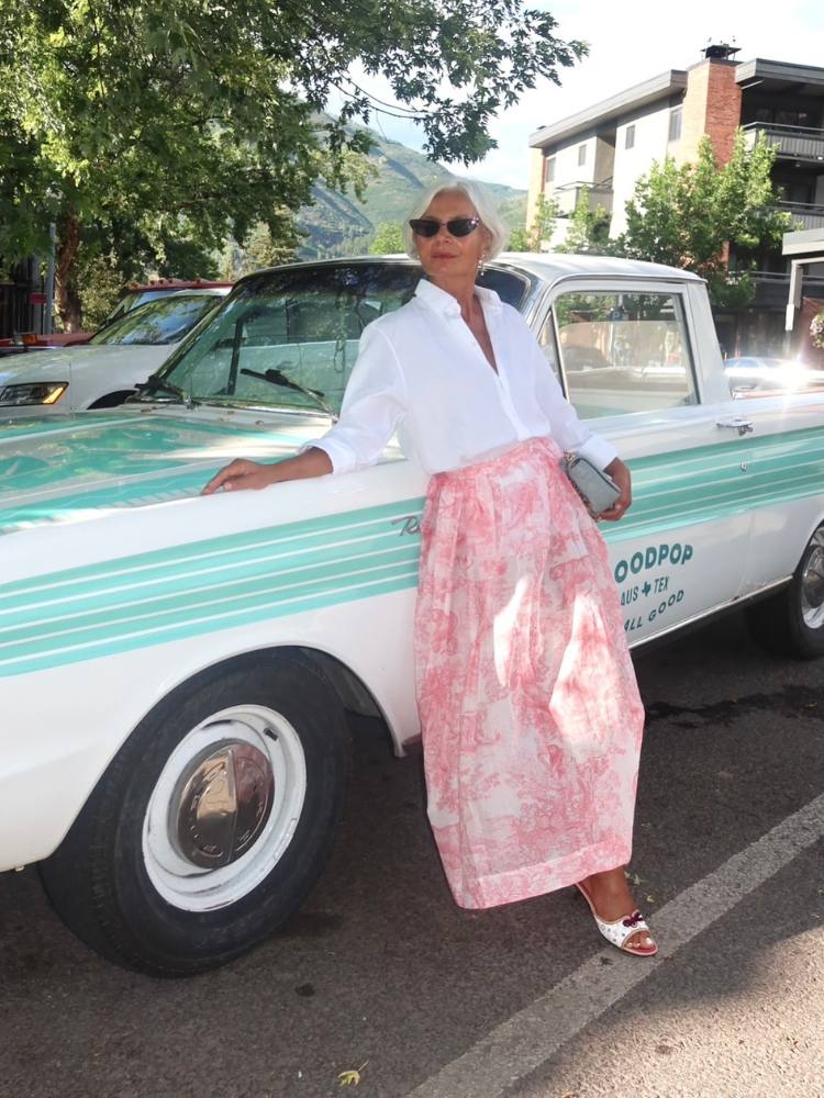 Senhora de pele clara apoiada em carro antigo branco e verde, usando camisa branca e saia longa rosa 