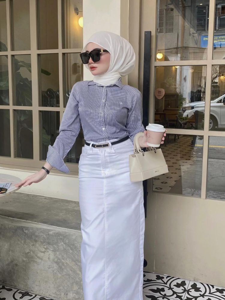 Mulher de pele clara usando hijab branco, camisa escura, saia longa branca, cinto marrom e bolsa bege