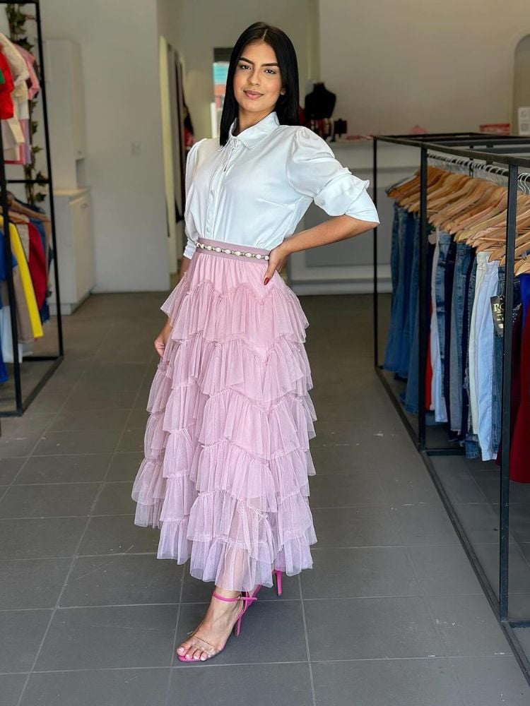 A imagem apresenta uma jovem em um ambiente de loja de roupas, posando com um look elegante e moderno. Ela veste uma camisa branca de botões com mangas curtas, que tem um corte clássico e sofisticado. Abaixo da camisa, ela usa uma saia midi em tom de rosa claro, com camadas de babados que conferem volume e movimento à peça. A saia é adornada com um cinto branco que possui detalhes em strass, adicionando um toque de brilho ao visual. Nos pés, ela calça sandálias de salto alto rosa, que complementam a saia e adicionam um ar feminino. O cabelo está solto e bem arrumado, e a expressão da jovem é confiante e descontraída. O fundo da imagem é neutro, com araras de roupas penduradas, destacando ainda mais o look da modelo.