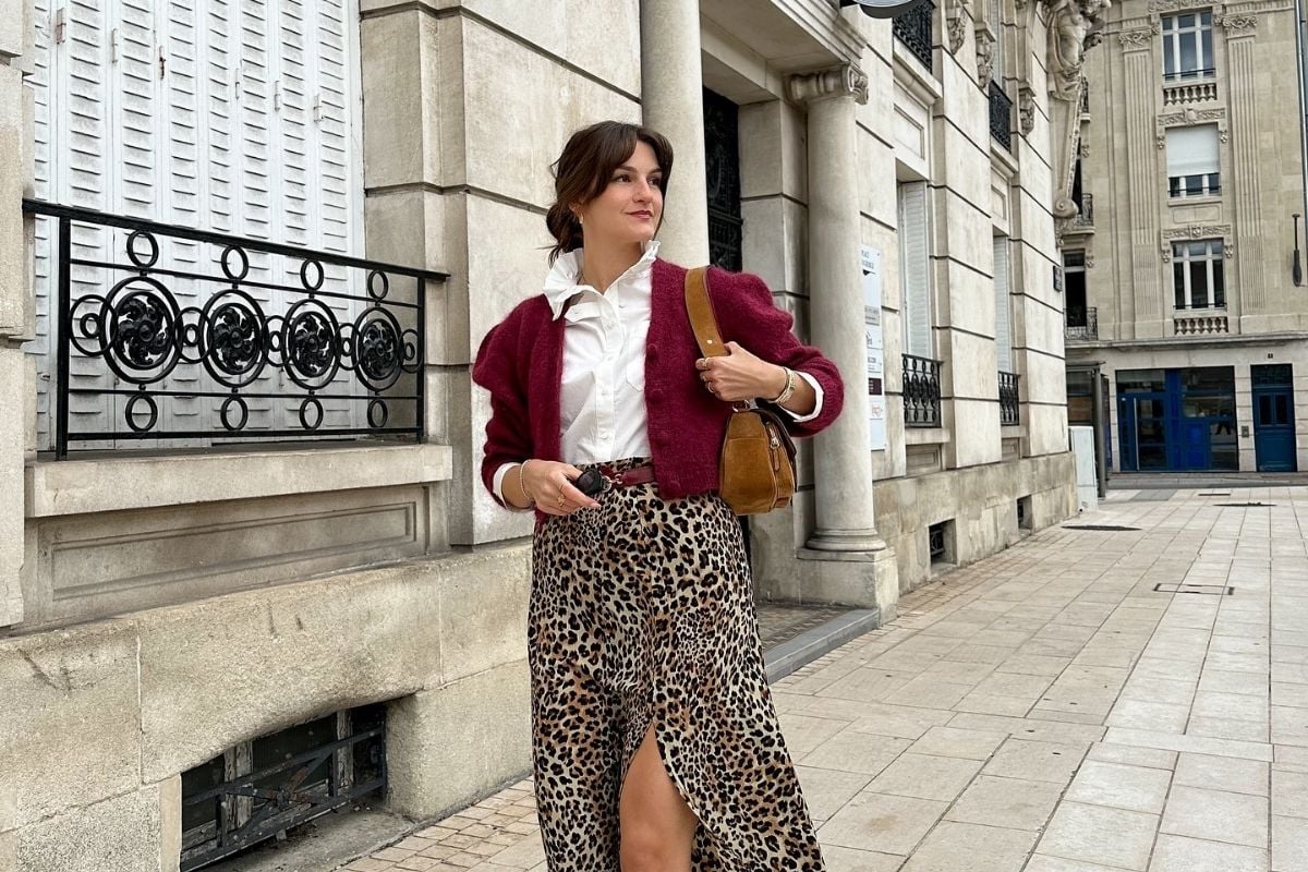 Mulher de pele clara em calçada, aparentemente europeia, usando camisa branca social com babados, casaco curto vinho, saia animal print onça com fenda e bolsa vinho