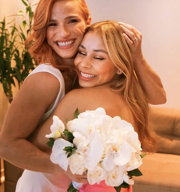 A imagem mostra duas mulheres sorridentes abraçadas. A mulher à esquerda tem cabelo ruivo e veste branco, enquanto a mulher à direita tem cabelo loiro e segura um buquê de flores brancas. Ambas parecem muito felizes, compartilhando um momento de alegria e proximidade. Ao fundo, há uma planta e uma parede clara.