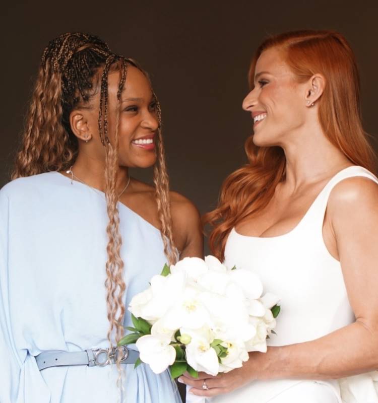 A imagem mostra duas mulheres sorridentes olhando uma para a outra. A mulher à esquerda tem cabelo trançado e veste um vestido azul-claro. A mulher à direita tem cabelo ruivo e veste um vestido branco, segurando um buquê de flores brancas. Ambas parecem felizes, compartilhando um momento de alegria e cumplicidade.