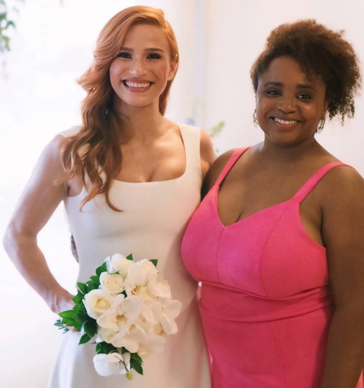 A imagem mostra duas mulheres sorridentes. A mulher à esquerda tem cabelo ruivo, veste um vestido branco e segura um buquê de flores brancas. A mulher à direita tem cabelo encaracolado, veste um vestido rosa e sorri. Ambas parecem felizes e posam juntas em um ambiente iluminado e claro, transmitindo um sentimento de alegria.