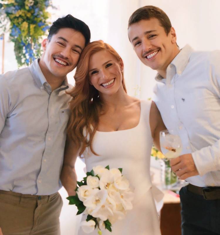 A imagem mostra três pessoas sorridentes em um ambiente claro. A mulher ao centro tem cabelo ruivo, veste um vestido branco e segura um buquê de flores brancas. Os dois homens ao seu lado vestem camisas sociais claras e sorriem. Todos parecem felizes, compartilhando um momento especial juntos.