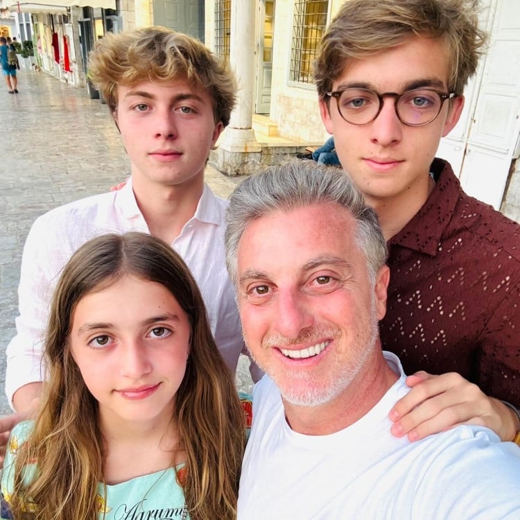 A imagem mostra quatro pessoas sorrindo juntas em um ambiente externo. Na frente, à direita, há um homem de cabelos grisalhos curtos, vestindo uma camiseta branca. À esquerda, uma menina de cabelos longos e castanhos, usando uma blusa estampada. Atrás, dois jovens: o da esquerda tem cabelo loiro encaracolado e veste uma camisa clara, enquanto o da direita usa óculos e uma camisa marrom. O fundo é composto por uma calçada de pedra e edifícios com colunas e janelas. Todos parecem estar felizes e próximos.