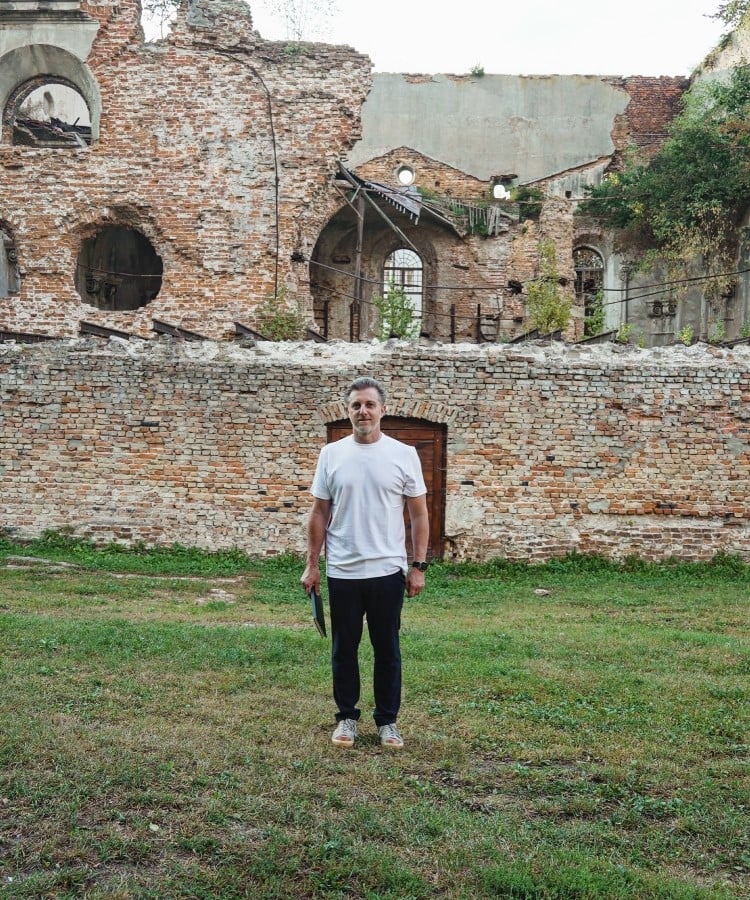 A imagem mostra Luciano Huck de pé em frente a uma parede de tijolos antigos e desgastados, que parece ser uma ruína histórica. Ele está vestindo uma camiseta branca e calças escuras, segurando algo na mão direita. O chão é coberto por grama verde, e há algumas árvores e vegetação ao fundo, além de janelas e arcos na estrutura em ruínas. O cenário transmite uma sensação de tranquilidade e história.