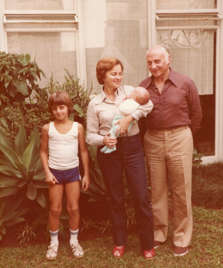 Na foto, há quatro pessoas em um jardim. À esquerda, Luciano Huck criança de cabelo castanho, vestindo uma camiseta branca e shorts azuis, está em pé. Ao lado, sua mãe de cabelo castanho curto, usando uma blusa clara e calças escuras, segura um bebê nos braços. O bebê está vestido de azul. À direita, seu pai, um homem mais velho, de cabelo grisalho, veste uma camisa marrom e calças bege. Todos estão sorrindo. Atrás deles, há plantas verdes e uma parede com janelas. O ambiente parece ser tranquilo e familiar.