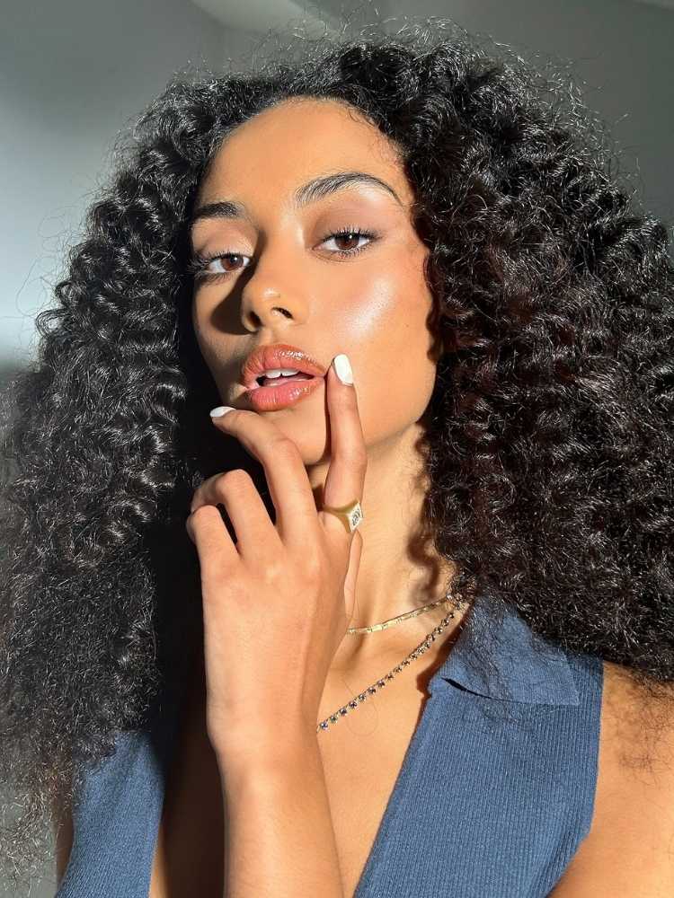 Mulher de pele morena e cabelo cacheado usando make suave, com sombra marrom clara, gloss e iluminador 