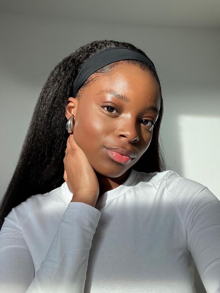 Mulher de pele negra usando blusa de manga longa branca, faixa preta no cabelo e maquiagem bem leve e fresca, com lábios corados de gloss, corretivo e blush