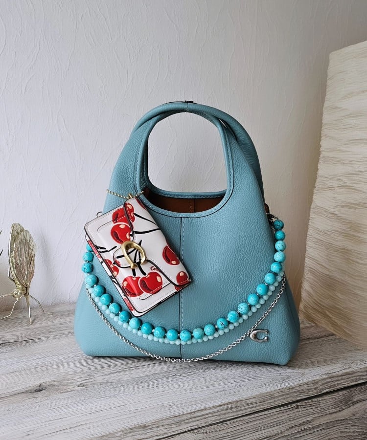 Uma bolsa azul claro com design curvo está adornada com um colar de contas turquesa e um lenço floral. O estilo maximalista é evidente, com acessórios que enriquecem o visual. As tendências em bolsas para 2025 destacam a personalização e o uso de elementos decorativos, trazendo vivacidade e individualidade às peças.