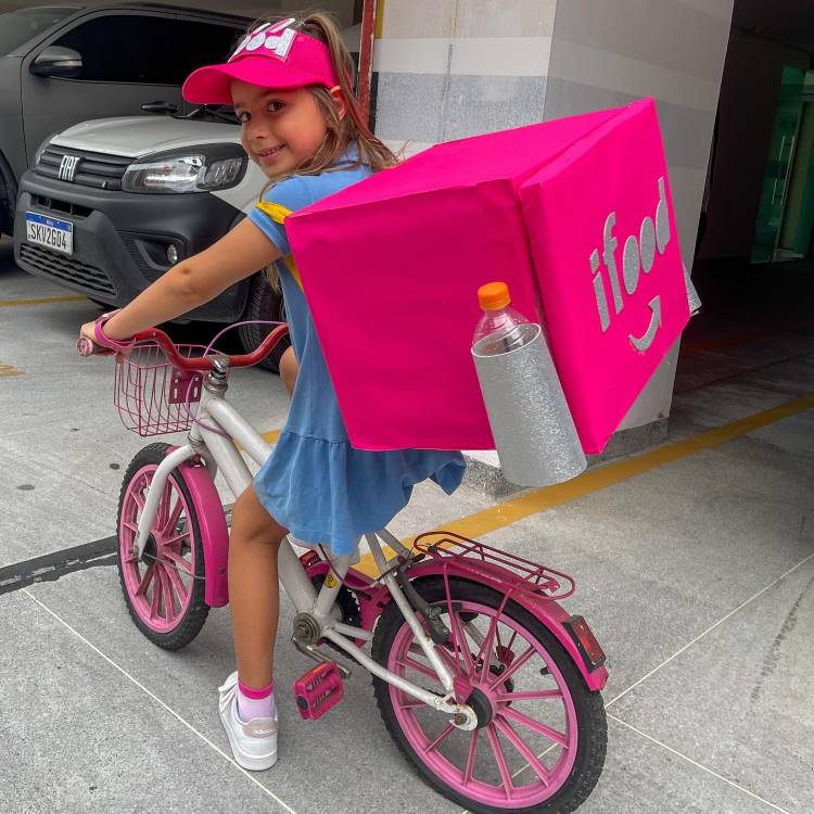 A imagem mostra uma menina sorridente em uma bicicleta rosa e branca, usando uma mochila inusitada em forma de caixa rosa brilhante. A mochila se assemelha a uma caixa de entrega de comida, com o logotipo "ifood" visível. A criança veste um vestido azul, boné rosa e tênis brancos. Ela está em uma área que parece ser um estacionamento, com um carro cinza ao fundo.