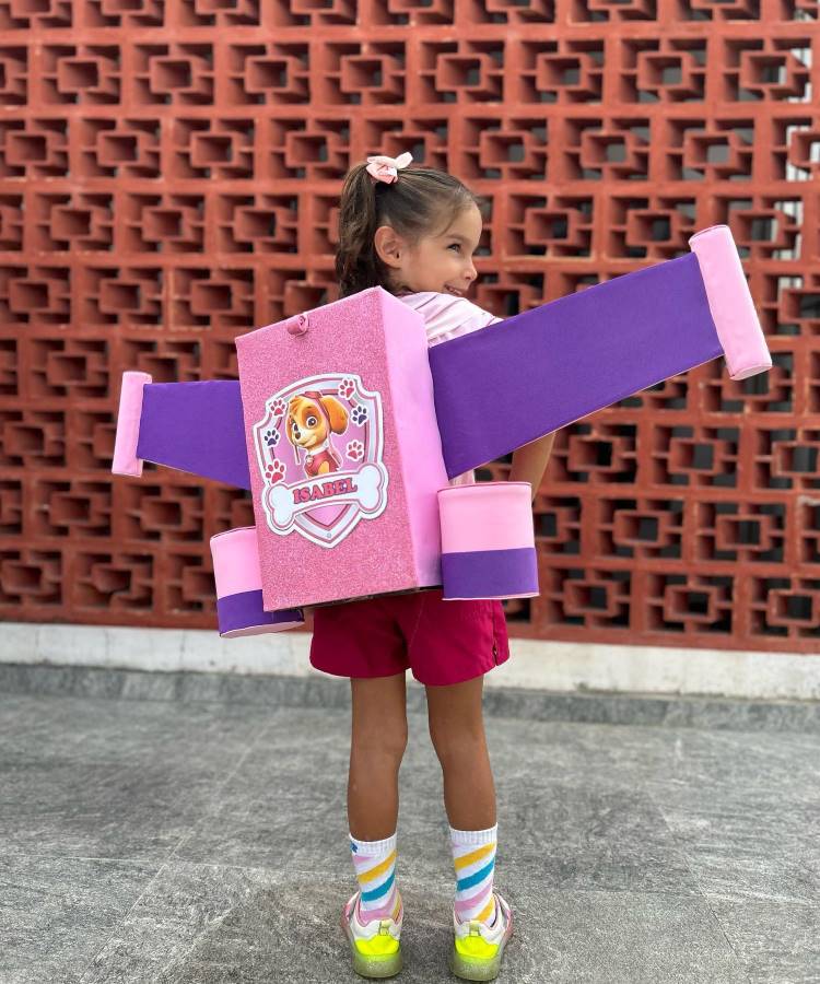 A imagem mostra uma criança usando uma mochila inusitada e criativa. A mochila tem formato de avião, com asas roxas e corpo rosa brilhante. Na frente, há um desenho da personagem Skye da Patrulha Canina. A criança veste shorts vermelhos, meias coloridas e tênis amarelos. Ela está em frente a uma parede de tijolos vazados vermelhos, olhando para o lado com um sorriso.