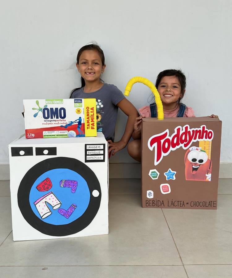 A imagem mostra duas crianças sorridentes com mochilas criativas feitas de caixas de papelão. À esquerda, uma menina carrega uma mochila que simula uma máquina de lavar roupas da marca OMO. À direita, uma criança menor usa uma mochila que representa uma caixa de Toddynho, bebida láctea de chocolate. As mochilas são coloridas e detalhadas, imitando fielmente as embalagens dos produtos.