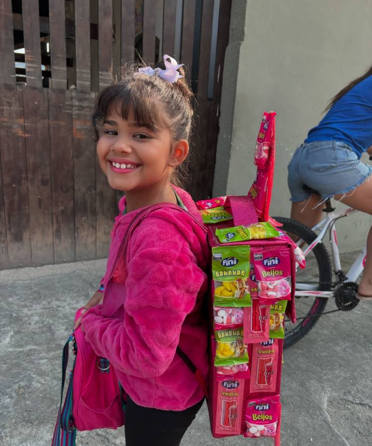 A imagem mostra uma menina sorridente com uma mochila maluca feita de embalagens de doces Fini. Ela veste um casaco rosa e tem um laço no cabelo. A criança está em frente a uma cerca de madeira, com parte de uma bicicleta visível. Sua mochila inusitada é composta por várias caixas de balas, criando um visual colorido e divertido que combina com seu sorriso radiante.