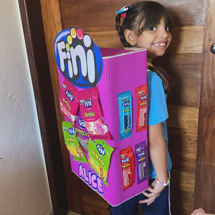 A imagem retrata uma menina sorridente usando uma mochila maluca e criativa feita para parecer uma caixa de doces Fini. Esta mochila inusitada é rosa vibrante, retangular e decorada com embalagens coloridas de diversos produtos Fini. A criança veste uma camiseta azul e tem um acessório no cabelo. Ela está posicionada em frente a uma porta de madeira, exibindo orgulhosamente sua mochila única.