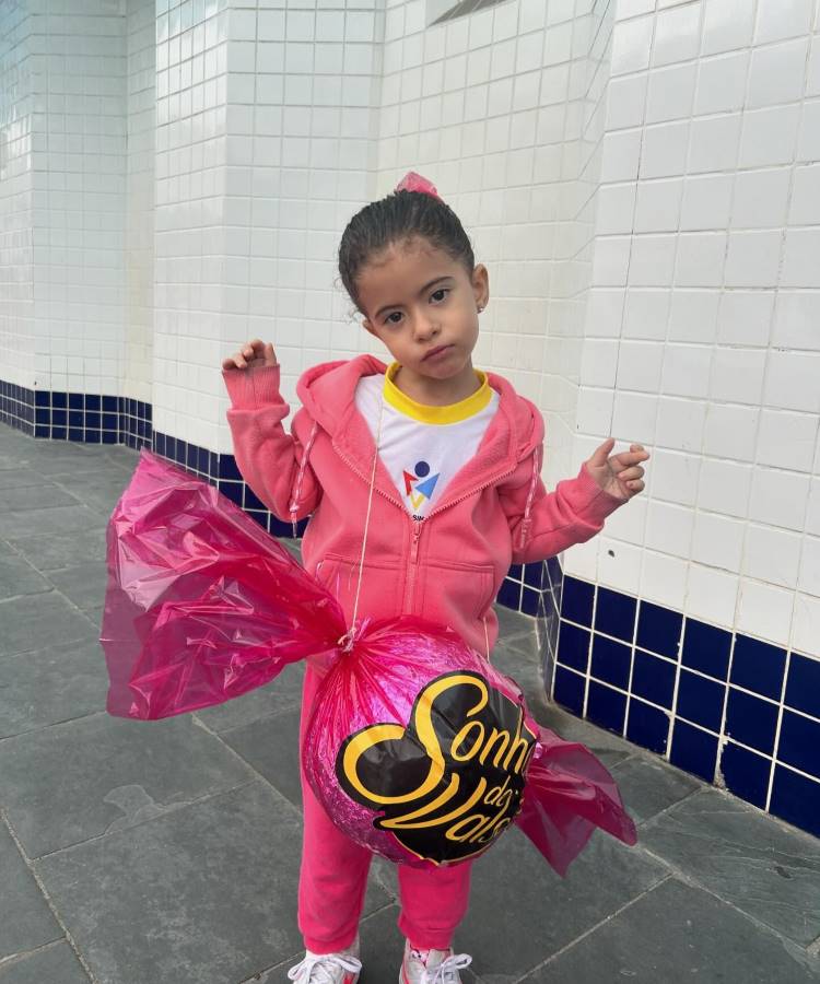 A imagem mostra uma menina vestida inteiramente de rosa, segurando uma mochila maluca em forma de bola de chocolate "Sonho de Valsa". Ela usa um conjunto esportivo rosa e um laço rosa no cabelo. A criança está em um ambiente com paredes de azulejos brancos e detalhes em azul escuro. Sua expressão é séria enquanto exibe sua mochila inusitada, que parece ser feita de material plástico brilhante.