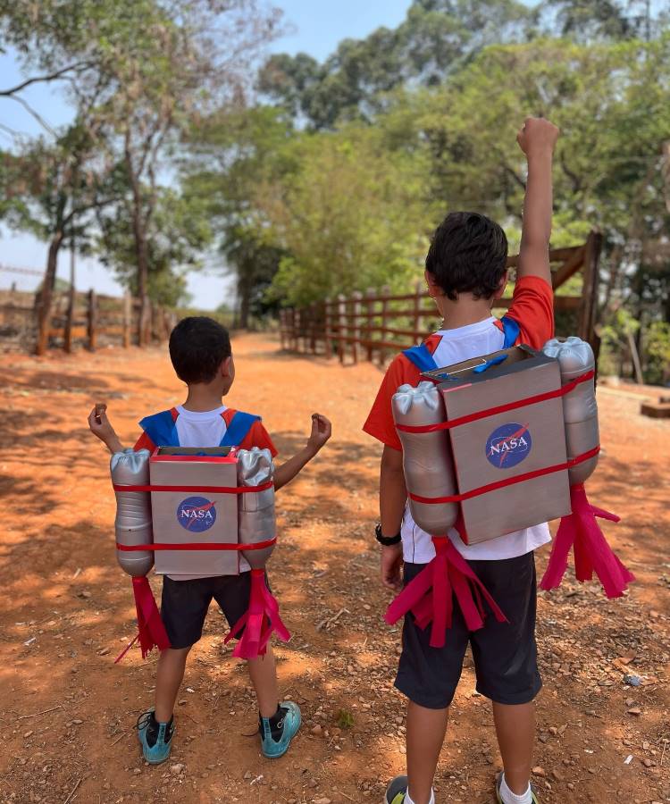 A imagem retrata dois meninos em um cenário rural, usando mochilas malucas inspiradas na NASA. As mochilas criativas simulam jetpacks espaciais, com o logotipo da NASA e "propulsores" vermelhos. Os garotos, vestindo camisetas brancas e shorts escuros, estão em uma estrada de terra com árvores ao fundo. Eles erguem os braços em um gesto triunfante, como se estivessem prestes a decolar em uma aventura espacial imaginária.