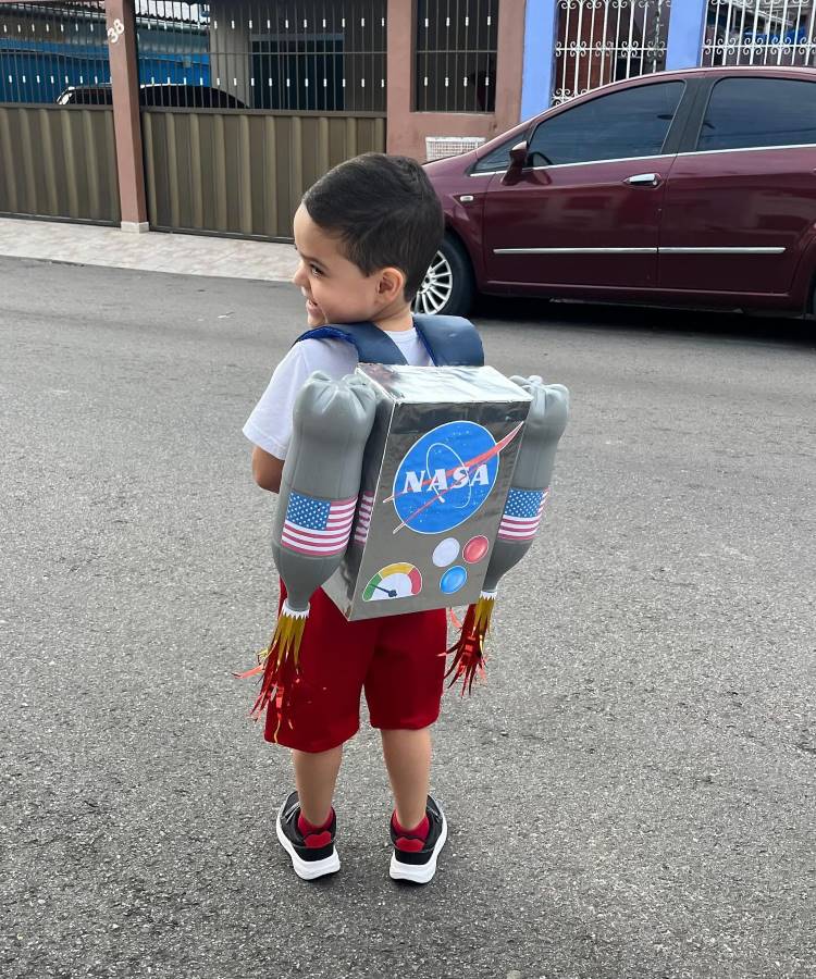 A imagem mostra um menino usando uma mochila maluca inspirada na NASA. Ele veste uma camiseta branca, shorts vermelhos e tênis. A mochila criativa simula um foguete espacial, com o logotipo da NASA, bandeiras americanas e "propulsores" feitos de garrafas plásticas. O garoto está em uma rua, com um carro e grades ao fundo, aparentando estar pronto para uma aventura espacial imaginária.