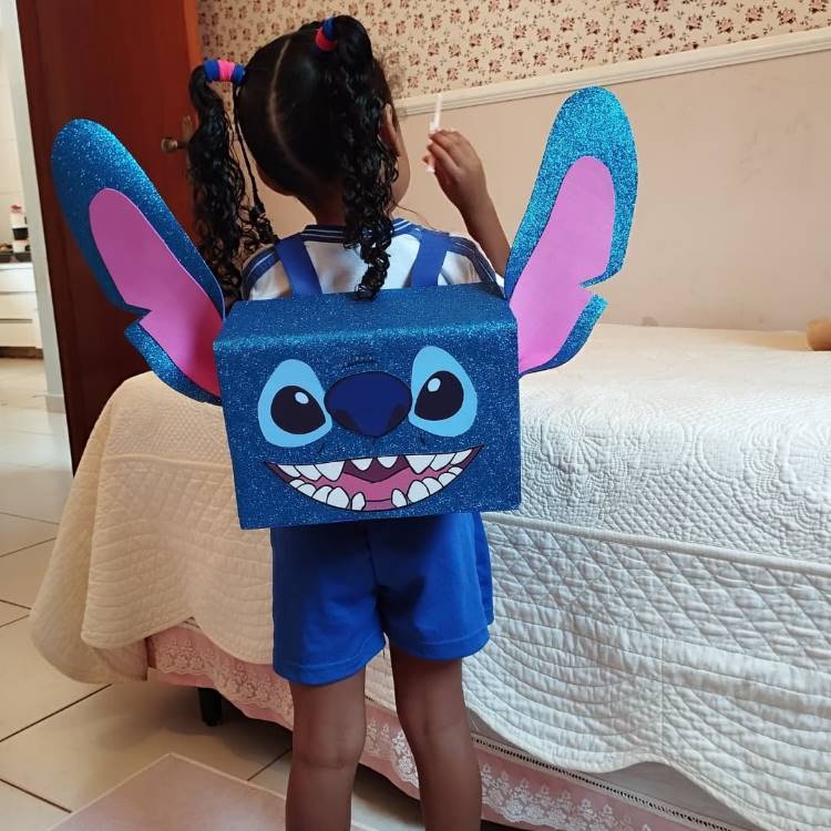 A imagem mostra uma criança usando uma mochila criativa inspirada no personagem Stitch, da Disney. A mochila tem formato retangular, é azul brilhante com glitter, e possui grandes orelhas características do Stitch. O rosto do personagem está representado na frente, com olhos grandes e um sorriso cheio de dentes. A criança veste shorts azuis e está de costas para a câmera, em um quarto com cama e papel de parede floral.