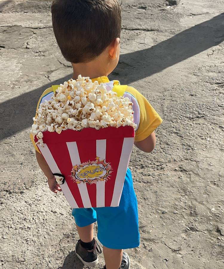 A imagem mostra uma criança de costas usando uma mochila maluca em forma de balde de pipoca. A mochila é vermelha e branca, com listras verticais e repleta de pipoca real. A criança veste uma camiseta amarela e shorts azuis, caminhando em uma superfície de concreto rachado. A sombra da criança é visível no chão, indicando um dia ensolarado.