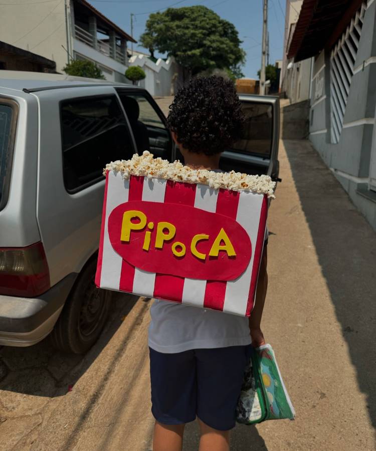 A imagem mostra uma criança de costas carregando uma mochila maluca em forma de caixa de pipoca gigante. A mochila é vermelha e branca com listras verticais, exibindo a palavra "PIPOCA" em amarelo. A criança está em uma rua residencial, próxima a um carro estacionado. Essa mochila criativa e inusitada certamente chama a atenção, transformando um item cotidiano em algo divertido e único.