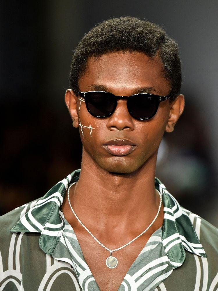 Zoom em homem de pele negra usando óculos de sol, linhas brancas no rosto e camisa verde estampada com orelha do Sansão 