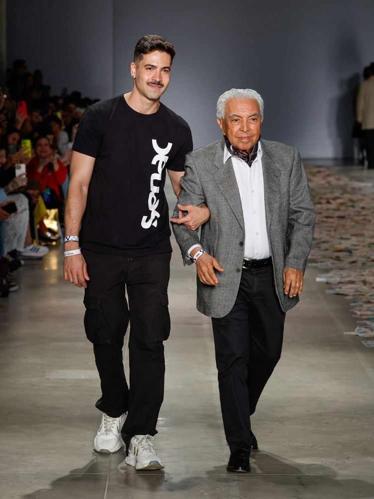 Rafael Caetano usando blusa preta do Senac e calça preta de braços dados com Mauricio de Souza em passarela da SPFW. O cartunista usa blazer cinza, camisa branca e calça preta