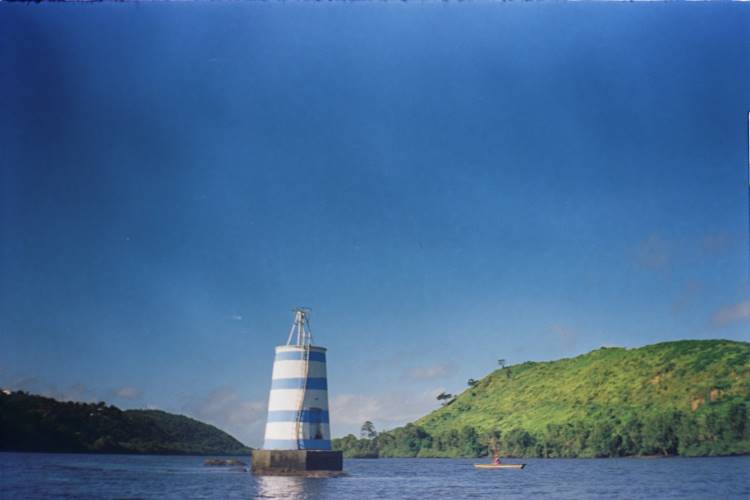 No Rio Paraguaçu, um farol listrado em azul e branco se destaca sob um céu límpido. As águas tranquilas refletem a luz suave, enquanto colinas verdes emolduram a cena ao fundo. Essa paisagem serena e vibrante inspira a nova coleção da Handred, capturando a essência natural e cultural da Bahia.