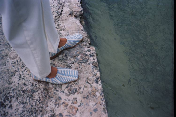 A imagem mostra uma pessoa à beira do Rio Paraguaçu, vestindo calças brancas e sapatos listrados em azul e branco. A borda de pedra contrasta com a água verde do rio, capturando a essência natural e tranquila da Bahia. Essa cena reflete a inspiração para a nova coleção da marca Handred.