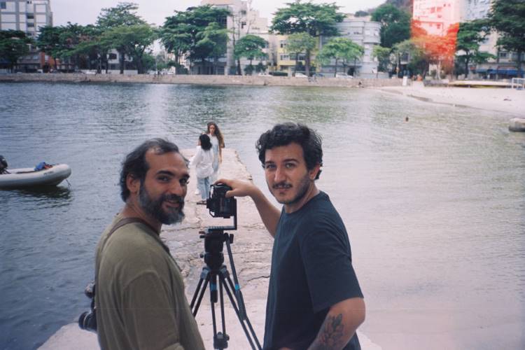 A imagem mostra dois homens próximos a uma câmera montada em um tripé, à beira do Rio Paraguaçu. Ao fundo, uma mulher caminha sobre um píer de pedra que se estende sobre as águas calmas. Árvores e prédios compõem a paisagem urbana ao redor, criando um contraste entre natureza e cidade.