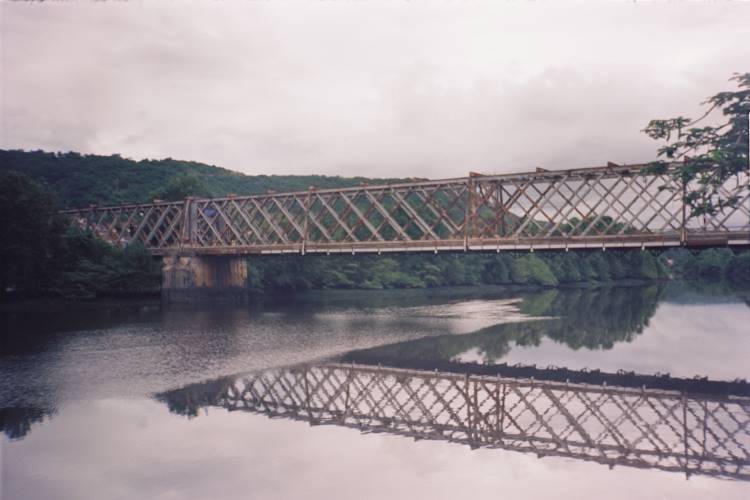 A imagem mostra uma ponte de estrutura metálica atravessando o Rio Paraguaçu, refletida nas águas calmas abaixo. Cercada por vegetação densa e colinas verdes, a cena transmite tranquilidade e beleza natural. O céu nublado adiciona um tom sereno à paisagem, destacando a integração harmoniosa entre a engenharia humana e o ambiente natural.