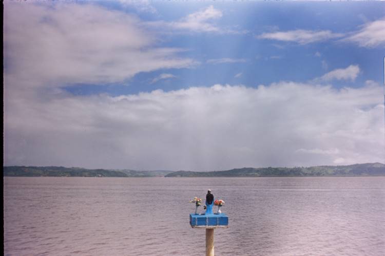 Sob um céu parcialmente nublado, o Rio Paraguaçu se estende em uma vasta superfície de águas tranquilas. No centro da imagem, uma estátua azul sobre uma plataforma eleva-se do rio, adornada com flores coloridas. Ao fundo, margens verdes e colinas suaves completam a paisagem serena e contemplativa.