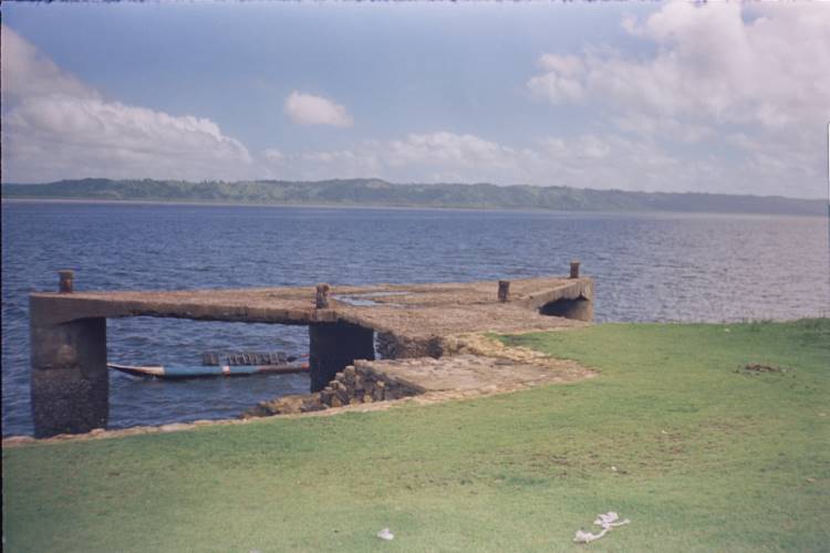 O Rio Paraguaçu se estende sob um céu azul, com um píer de pedra avançando sobre suas águas calmas. A vegetação verde das margens complementa a paisagem serena e natural. Essa cena inspira a nova coleção da Handred, capturando a essência tranquila e a beleza rústica da Bahia, refletindo elementos de natureza e arquitetura.